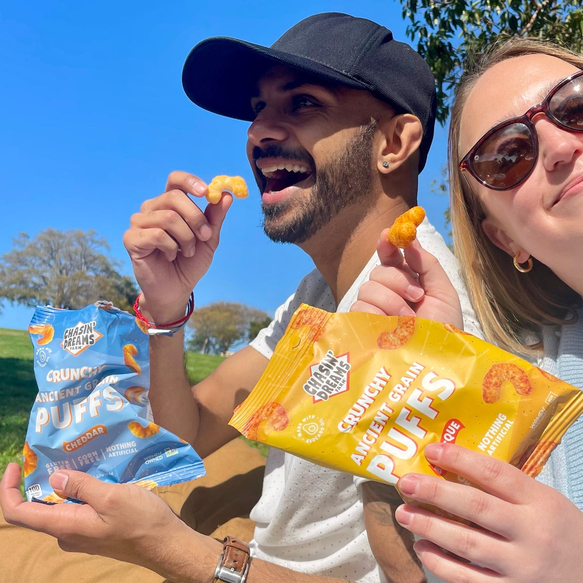 Man wearing baseball hat eating a bag of cheddar puffs, woman wearing sunglasses eating a bag of barbeque puffs.