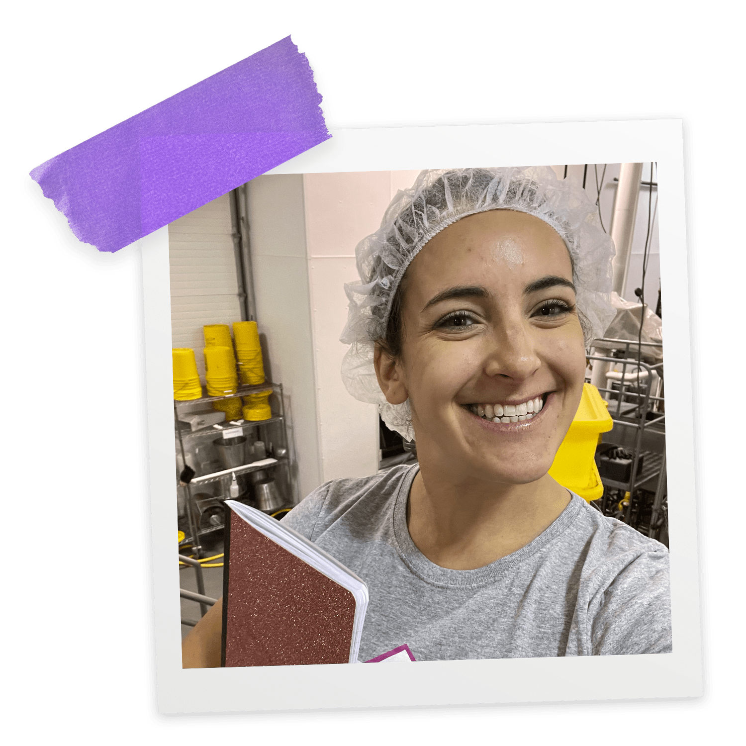 A woman stands in a manufacturing plant holding a notebook in one hand, smiling and wearing a hair net!